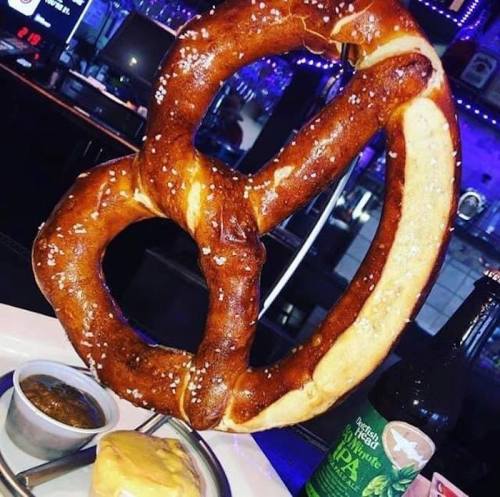 A large, twisted pretzel with salt, served with dipping sauce and a beer in the background.