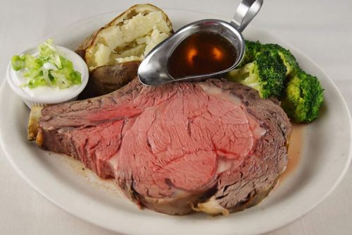 A plate featuring a large slice of prime rib, baked potato, broccoli, and a small bowl of sour cream.