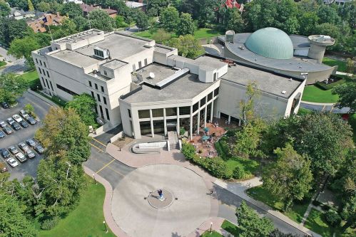 Aerial view of a modern building with a dome, surrounded by greenery and a circular plaza, with parked cars nearby.