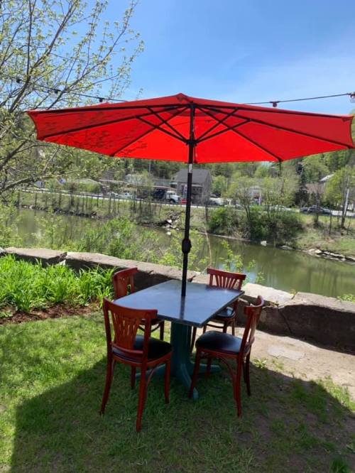 A table with red umbrella and chairs by a river, surrounded by greenery and a clear blue sky.