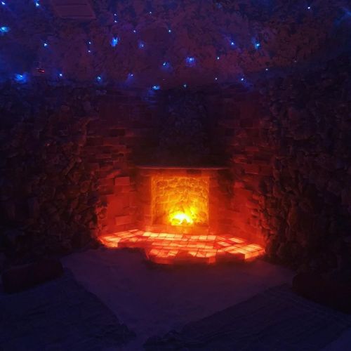 A cozy, dimly lit room with a glowing salt brick fireplace and blue ambient lights on the walls.