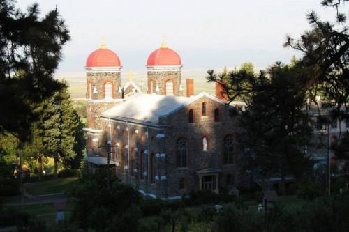 Historic stone building with red domes, surrounded by trees and greenery, under soft sunlight.