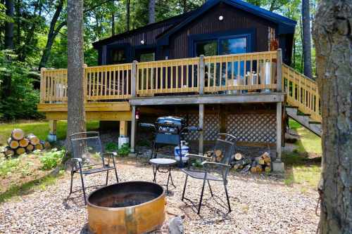A cozy cabin with a wooden deck, surrounded by trees, featuring a grill and fire pit with chairs in the foreground.