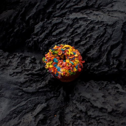 A colorful donut topped with vibrant sprinkles resting on a textured black surface.
