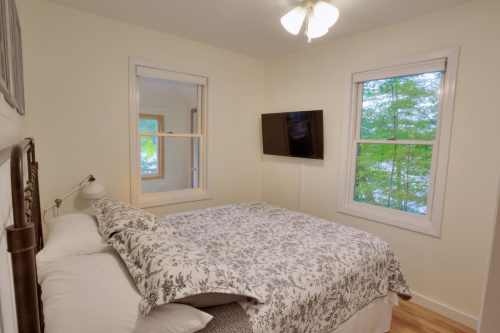 Cozy bedroom with a floral bedspread, two windows, and a wall-mounted TV, surrounded by greenery.