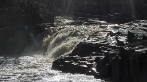 A waterfall cascades over rocky terrain, surrounded by trees, with sunlight reflecting off the water's surface.