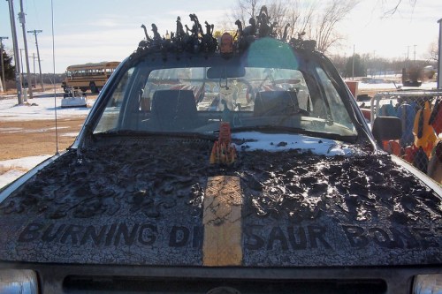 A burnt-out car with dinosaur figures on the roof, featuring the words "BURNING DINOSAUR POINT" on the hood.