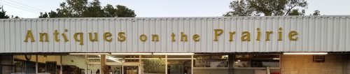 Sign for "Antiques on the Prairie" displayed on a storefront with a clear sky in the background.