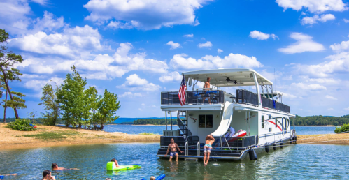 A houseboat anchored near a sandy shore, with people swimming and relaxing in the water under a blue sky.