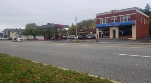 A gas station and a bakery storefront along a quiet road, with trees and buildings in the background under a cloudy sky.