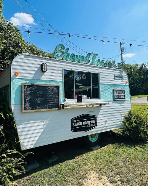 A vintage food truck with a teal exterior, serving snacks, surrounded by greenery and under a clear blue sky.