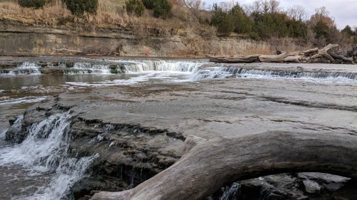 A serene river scene featuring cascading waterfalls over rocky ledges, surrounded by trees and a log on the shore.