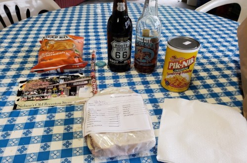 A table with a blue checkered tablecloth featuring snacks, drinks, a sandwich, and a postcard.