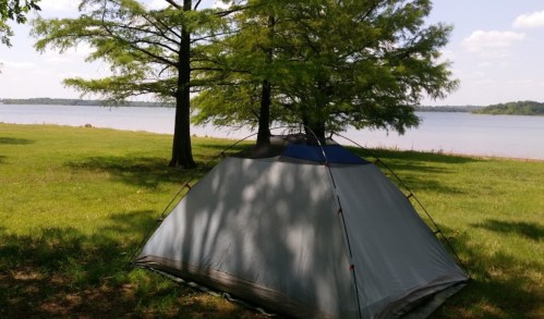 A tent set up on grassy land near a calm lake, surrounded by trees under a clear blue sky.