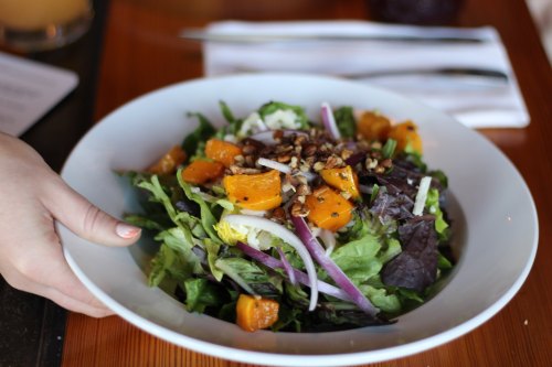 A hand holds a bowl of mixed greens salad topped with roasted butternut squash, red onion, and pecans.