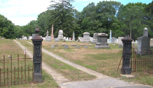 A cemetery entrance with wrought iron gates, leading to a grassy area filled with various gravestones and trees.