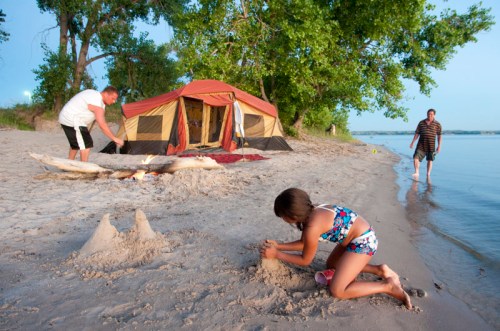 A girl builds a sandcastle on the beach while two adults set up a tent nearby. Trees and water are in the background.