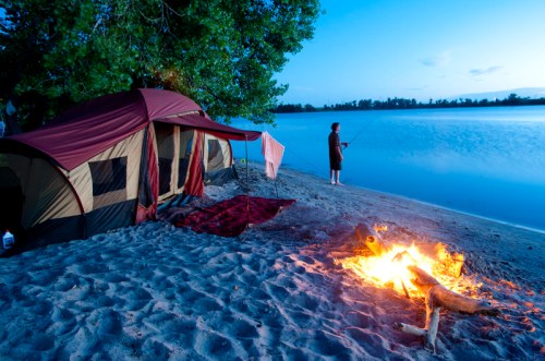 A person fishing by a calm lake at dusk, with a tent and a campfire on the sandy shore.