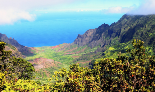 A vibrant landscape featuring lush green mountains descending to a blue ocean, under a partly cloudy sky.
