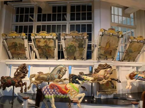 A display of colorful carousel horses in front of vintage painted panels in a bright, airy room.