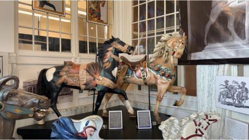 Two intricately painted carousel horses displayed in a museum, with additional horse sculptures in the background.