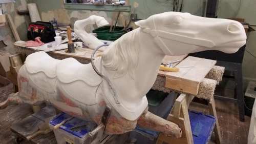 A partially restored carousel horse sculpture on a workbench, surrounded by tools and materials.