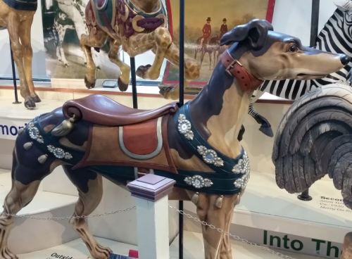 A decorative wooden greyhound statue with ornate detailing and a saddle, displayed in a museum setting.