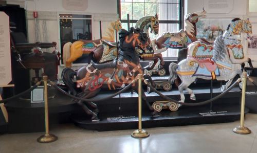 A display of colorful carousel horses in a museum setting, with ropes and stanchions for protection.