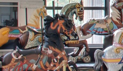 Colorful carousel horses in a display, featuring intricate designs and details, set against a bright background.