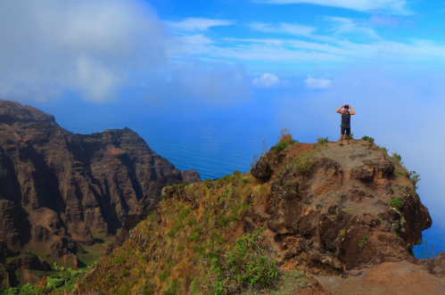 A person stands on a rocky cliff overlooking a vast ocean and dramatic mountain landscape under a blue sky.