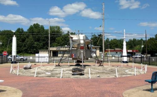 A replica of a lunar lander surrounded by rocks and markers, set in a park with blue skies and trees in the background.