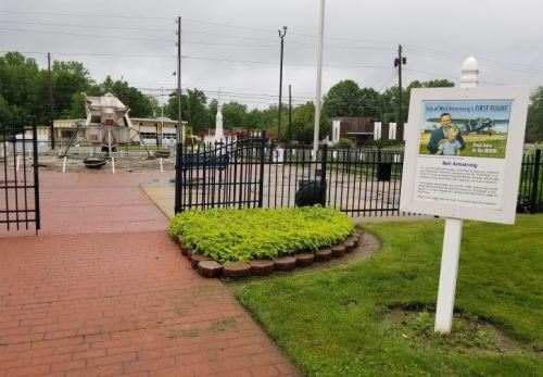 Entrance to a park featuring a sign about Neil Armstrong's first flight, with a lunar module in the background.