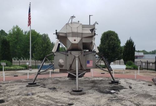 A replica of a lunar lander displayed outdoors, surrounded by trees and a flag in the background.