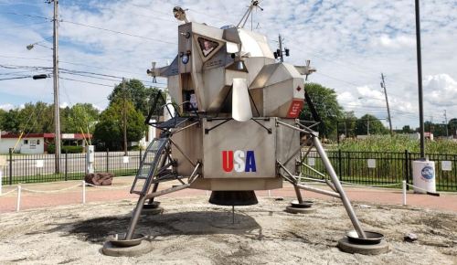 A replica of a lunar lander with "USA" displayed, set in a park with trees and a blue sky in the background.