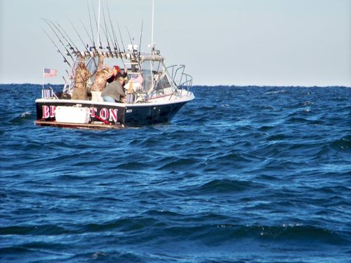 A fishing boat with multiple rods and flags, navigating through blue ocean waters on a clear day.