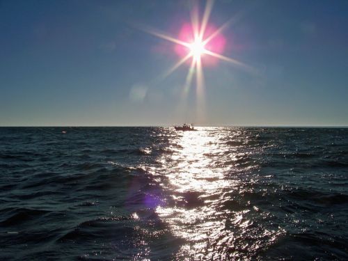 A bright sun shines over a shimmering ocean, with a small boat in the distance against a clear blue sky.