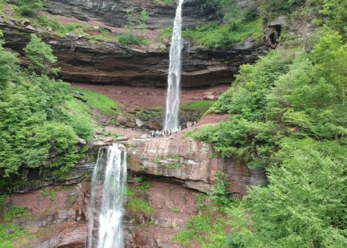 Two waterfalls cascade down rocky cliffs surrounded by lush greenery, with people visible at the base.
