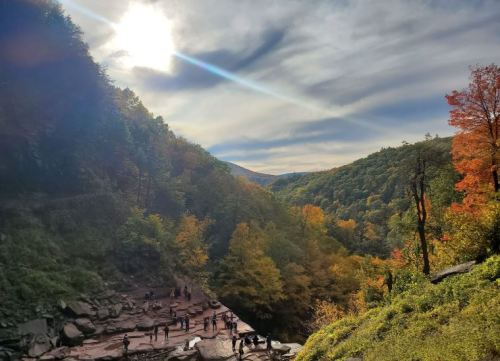 A scenic view of a valley with colorful autumn trees, a bright sun, and people exploring the rocky terrain below.