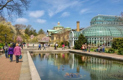 A vibrant park scene with a glass conservatory, blooming trees, and people walking along a pond under a blue sky.