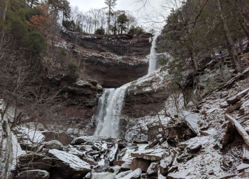 A snowy landscape featuring a tall waterfall cascading down rocky cliffs, surrounded by trees and winter scenery.