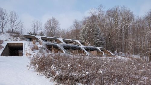 Snow-covered landscape featuring modern, bunker-like structures nestled in a wooded area.