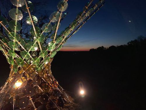 A sculpture made of colorful discs illuminated at dusk, with a sunset and dark landscape in the background.