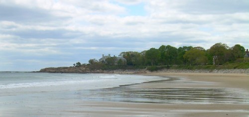 A serene beach scene with gentle waves, sandy shore, and a distant house surrounded by trees under a cloudy sky.