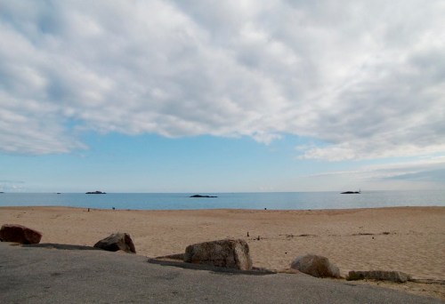 A serene beach scene with soft sand, calm water, and a cloudy sky, featuring distant rocks on the horizon.