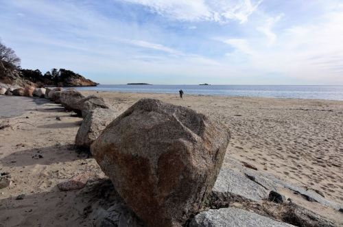 A rocky beach with large boulders in the foreground and a calm sea under a blue sky in the background.
