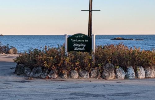 A sign reading "Welcome to Singing Beach" near a calm ocean, surrounded by rocks and coastal vegetation.