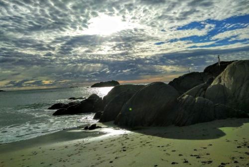 A serene beach scene with rocky formations, gentle waves, and a cloudy sky reflecting sunlight at sunset.