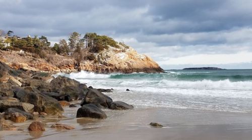 A rocky beach with gentle waves, under a cloudy sky and a coastal cliff in the background.