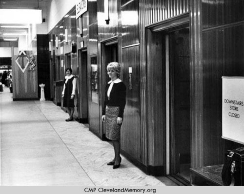 Black and white photo of two women in a corridor with elevators, one standing and the other in the background.