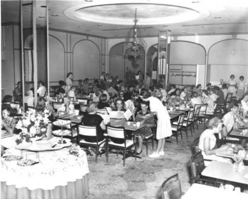 A busy dining room filled with people at tables, with waitstaff serving food in a vintage setting.